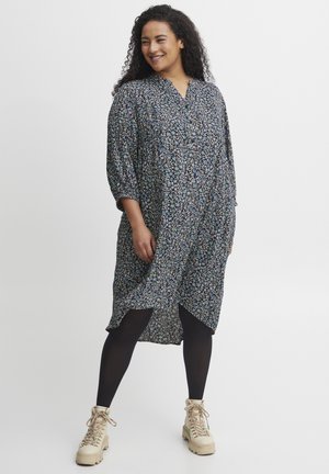 Blommönstrad klänning med V-ringning, långa ärmar och en loose fit. Mörk bakgrund med färgglada mönster. Bärs med svarta strumpbyxor och beige stövlar.