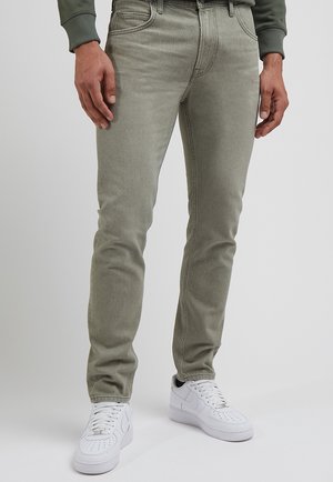 Homme portant un jean slim fit vert olive clair et des baskets blanches, debout devant un fond uni.