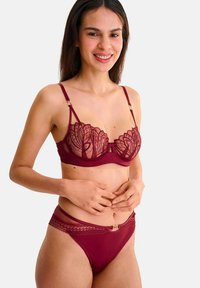 Soutien-gorge en dentelle bordeaux avec un motif floral et des bretelles ajustables, associé à un string avec des accents en dentelle assortis orné d'un emblème décoratif en or.