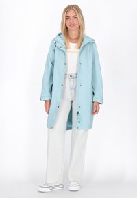 Gaiši zilā ūdensizturīgā lietusmantu ar kapuci, priekšējo rāvējslēdzēju un spiedpogām, uzvilkta pāri baltai krekla un gaišām džinsu biksēm. Gluda tekstūra.