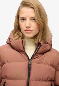 Chaqueta acolchada color rosa con cuello alto, cremallera negra y detalles de cordón, hecha de un material suave y ligero.