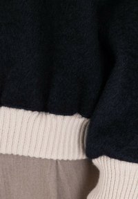 Indumento di lana nera con una superficie texturizzata, caratterizzato da un orlo a coste color crema. Il tessuto è morbido e contrasta con una sezione liscia beige sottostante.