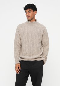 Bež pleteni turtleneck pulover, s teksturiranim uzorcima, dugim rukavima i opuštenim krojem, nosi se uz crne hlače.