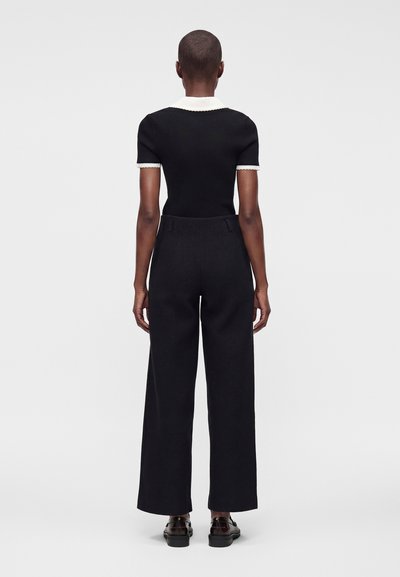 Personne aux cheveux courts vue de dos portant un haut noir à manches courtes avec col blanc et un pantalon noir à jambes larges avec des chaussures noires.