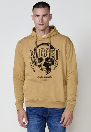 Sudadera marrón con un gráfico de calavera negra y el texto "RUGGED", con capucha con cordón y puños de canalé, combinada con jeans azules.