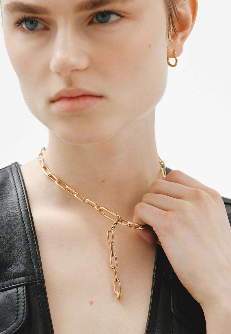 Collar de cadena de oro con eslabones alargados y un acento colgante. El modelo también lleva un pequeño pendiente de aro. Se puede ver una parte superior de cuero negro.