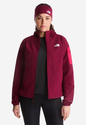 Ung kvinde iført en bourgogne lynlåstrøje og matchende pandebånd, begge med The North Face-logo, stående mod en hvid baggrund.