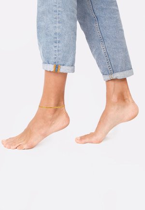 Cavigliera in oro appoggiata su una caviglia, abbinata a jeans in denim azzurro chiaro arrotolati. La posizione a piedi nudi mette in risalto la texture e i dettagli della pelle.
