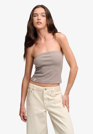 Jeune femme aux longs cheveux foncés porte un haut rayé marron et blanc sans bretelles et un pantalon large écru taille haute.