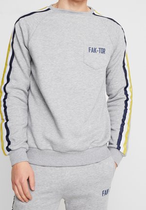 Mężczyzna ubrany w jasnoszarą bluzę i pasujące spodnie z granatowymi i żółtymi paskami na rękawach oraz napisem "FAK-TOR" na kieszeni piersiowej.