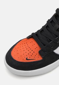 Sapatilha preta e laranja com parte superior em camurça, caixa dos dedos perfurada, atacadores pretos e sola de borracha branca com um pequeno logotipo da Nike.