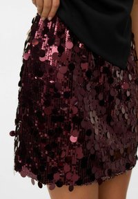 Krátká bordó sukně se sametovým povrchem, zdobená kulatými flitry uspořádanými těsně vedle sebe pro reflexní efekt. Okraje jsou bez švů.
