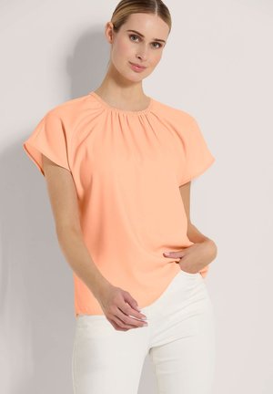 Vrouw met licht haar die een losse perzikkleurige korte mouwen blouse en witte broek draagt, staand met één hand in de zak tegen een effen achtergrond.