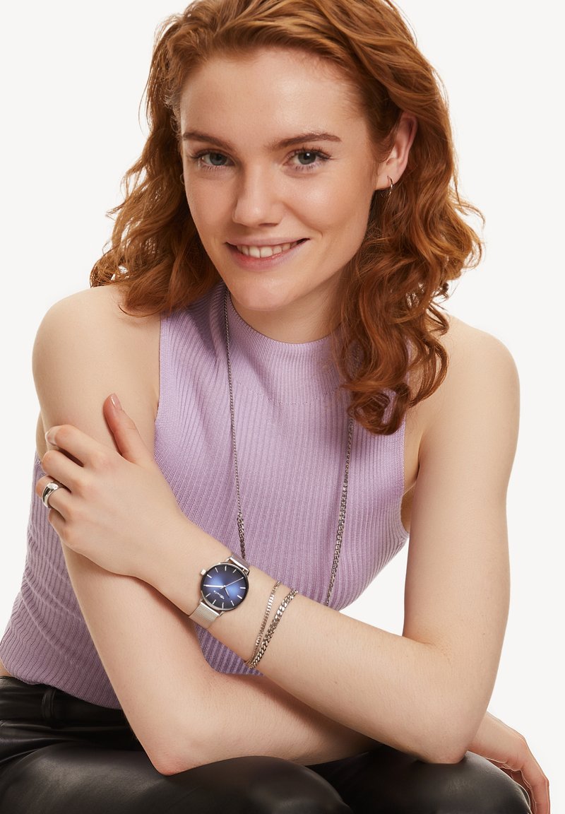 Orologio con cinturino in rete argento, quadrante circolare blu, indicatori delle ore argentati e design minimalista. Modella che indossa un top senza maniche color lilla e braccialetti.
