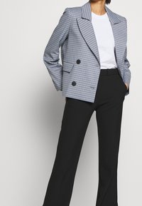 Dubbelknäppt kavaj i ljusblått och vitt rutig mönster, med slag och svarta knappar, i kombination med svarta byxor och en vit skjorta.