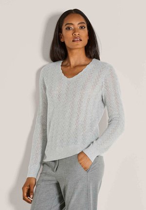 Frau in leicht grauem, gemustertem Strickpullover und grauen Hosen, steht mit einer Hand in der Tasche vor einem einfarbigen Hintergrund.