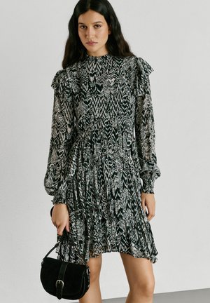 Kvinde med langt mørkt hår iført en sort-hvid mønstret kjole med lange ærmer, holder en lille sort håndtaske og står foran en ensfarvet baggrund.