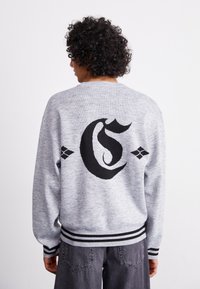 Grå strikket sweatshirt med et stort sort bogstav 'G' grafik på ryggen, samt sort-hvide stribede ribkantede manchetter og kant.