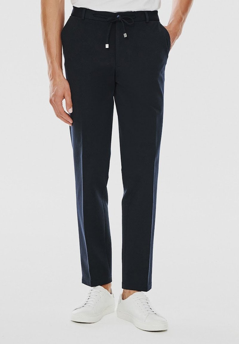 Granatowe spodnie z elastycznym paskiem, slim fit, ozdobione kwadratowymi metalowymi detalami. W połączeniu z białymi sneakersami. Gładka tekstura materiału.