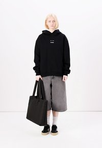 Black hoodie with front graphic and a relaxed fit, paired with gray shorts and a black tote bag. Model wearing black sneakers and white socks.