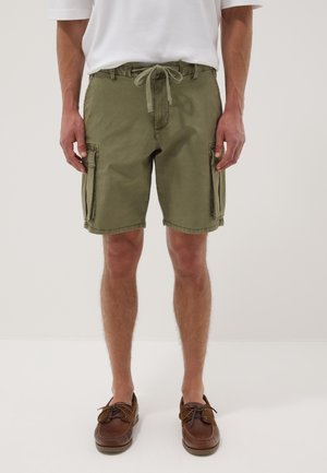 Mann trägt olivgrüne Cargo-Shorts mit Kordelzugbund und weißem T-Shirt, steht barfuß auf einfarbigem Hintergrund.