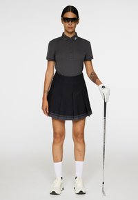 Vrouw in een donkere polo, zwarte geplooide rok, witte golfhandschoen, zonnebril en sneakers, die rechtop staat met een golfclub in de hand.