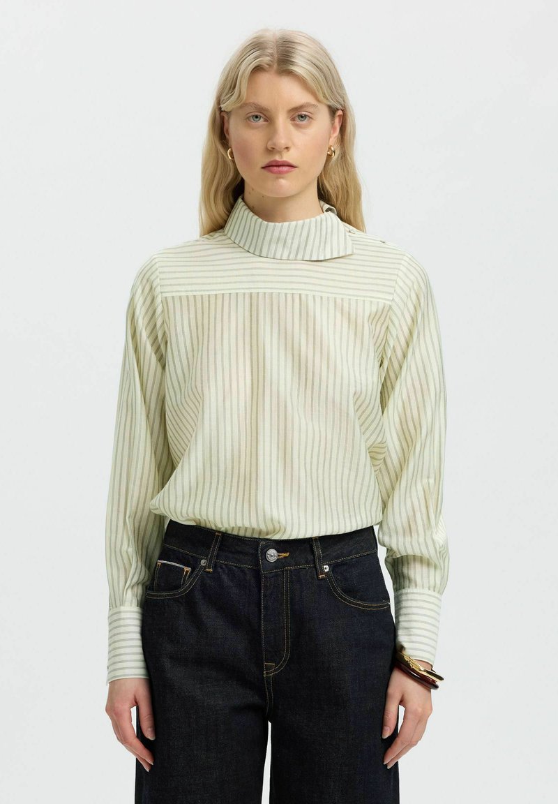 Femme aux longs cheveux blonds portant un chemisier clair à rayures et col montant, ainsi qu'un jean foncé, debout devant un fond blanc uni.