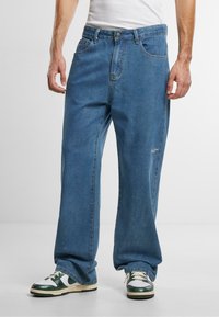 Jeans de mezclilla azul con un corte suelto, con cierre de botón en la parte delantera, cinco bolsillos y un logotipo sutil en el muslo. Llevados con zapatillas verdes y blancas.