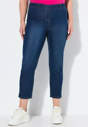 Woman wearing dark blue cropped denim jeans with elastic waistband and white platform sneakers against a plain white background.