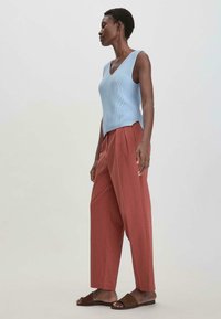 Vaaleansininen hihaton neulepaita, jossa on V-pääntie; rento tiilenpunainen leveälahkeinen housut; ruskeat liukupohjaiset sandaalit; sileät pinnat.