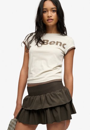 Joven con cabello largo y oscuro que lleva una camiseta beige con texto parcial y una falda oscura en capas sosteniendo un artículo de cuadros en la mano.