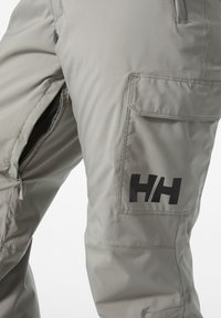 Hellgraue Hosen mit glatter Textur, ausgestattet mit einem großen schwarzen Logo, einem Seitenreißverschluss und einem Detail im Cargo-Stil auf der Rückseite.