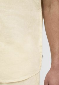 Tissu de chemise en lin jaune clair, présentant une finition texturée et une étiquette sur le côté. La chemise a une coupe droite avec un ourlet courbé.