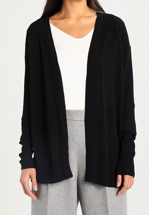 Femme portant un cardigan noir ample ouvert sur le devant, par-dessus un haut blanc, avec un pantalon gris taille haute à jambes larges.