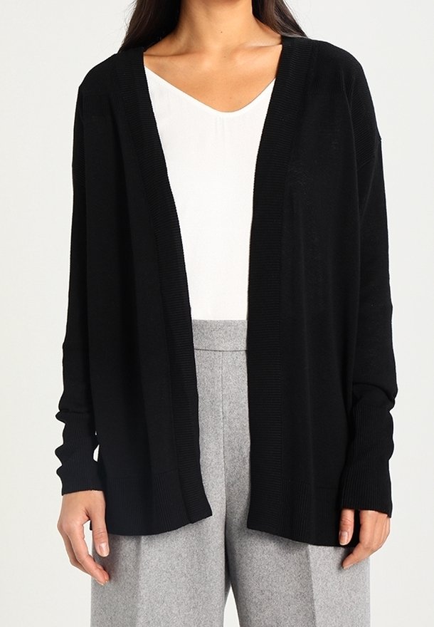 Femme portant un cardigan noir ample ouvert sur le devant, par-dessus un haut blanc, avec un pantalon gris taille haute à jambes larges.