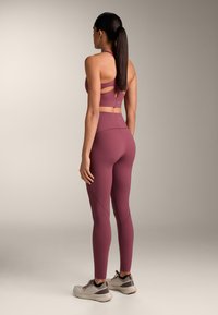 Donna con capelli lunghi e scuri vista di spalle, indossa un reggiseno sportivo color malva e leggings a vita alta abbinati, con scarpe da corsa beige.