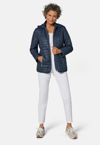 Veste matelassée bleu marine avec capuche, dotée de poches zippées et de détails côtelés. Portée avec un pantalon blanc et des baskets légères.
