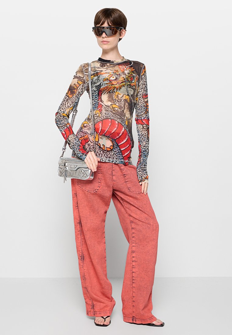 Langärmeliges, grafisches T-Shirt mit kühner Drachenmotivadaption, kombiniert mit lockeren, ausgebleichten roten weiten Hosen. Das Model trägt eine silberne Umhängetasche.