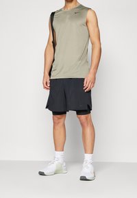 Szary bezrękawnik sportowy, czarne szorty z wewnętrzną podszewką, białe i jasnozielone sneakersy, zaprezentowane z torbą na ramię.