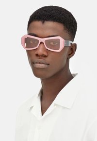 Occhiali da sole rosa oversize con montatura angolare, lenti semi-trasparenti e bracci neri a contrasto. Indossati con una camicia bianca.