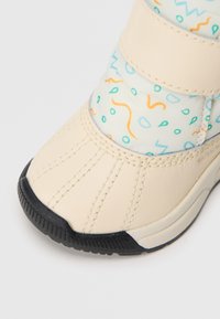 La bota presenta una parte superior de cuero crema, un eje textil estampado con diseños abstractos en azul y naranja, y una suela de goma negra texturizada.