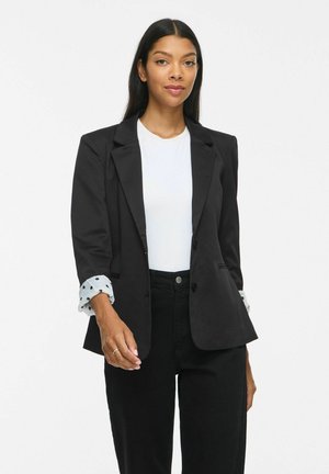 Femme aux longs cheveux foncés portant un blazer noir sur une chemise blanche et un pantalon noir, debout devant un fond blanc uni.
