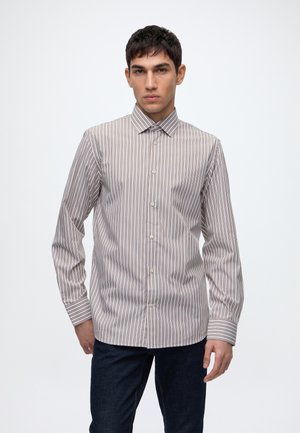 Homme portant une chemise à manches longues boutonnée avec des rayures verticales marron et blanches et un pantalon foncé, debout devant un fond uni.