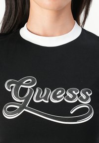 Juodas marškinėliai su išraiškingu baltu „Guess“ logotipu drąsiai rašytu šriftu, su baltu raišteliu aplink apykaklę.