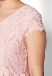 Närbild av en person som bär en rosa blommig spetsklänning med korta ärmar, som visar detaljerade spetsmönster på axel och överarm.