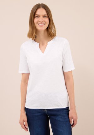Glunderende vrouw met schouderlang lichtbruin haar, gekleed in een witte kortmouwen V-hals top en donkerblauwe jeans, tegen een beige achtergrond.