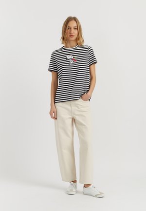 Donna che indossa una maglietta a righe bianche e nere con Snoopy che tiene delle ciliegie, pantaloni larghi color crema e sneakers bianche, in piedi su sfondo bianco.