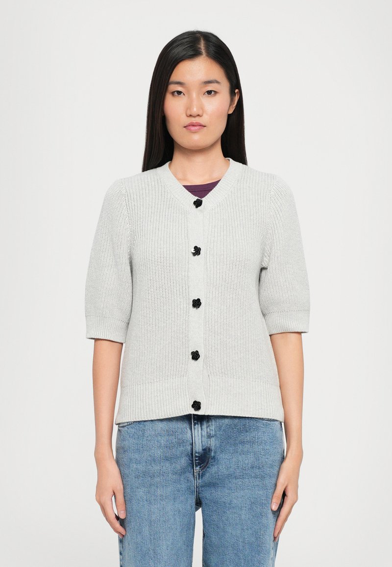 Lys grå strikket cardigan med V-hals, korte ermer og sorte knapdetaljer. Myk tekstur, tettsittende design og ribbestrikket kant.