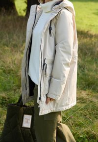 Veste matelassée beige avec capuche, dotée de fermetures à bascule et de poches latérales, portée par-dessus un pull blanc et un pantalon vert olive.