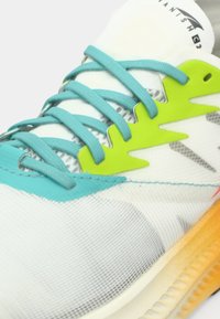 Zapatilla deportiva con parte superior de malla blanca, cordones turquesa, detalle verde con patrón en zigzag y suela con degradado de naranja a amarillo.